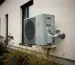A modern house with external heat pump unit installed next to the wall, captured on an overcast day in autumn. The outdoor heat exchanger shows condensation patterns on its metal fins, with pipes running from it into the building. In the foreground, a small garden area with some native plants. The scene has natural, neutral lighting typical of a cloudy day, with the heat pump appearing as an unassuming but integral part of the home's infrastructure. The outdoor unit is positioned against the house's light-colored exterior wall, highlighting the contrast between technology and domestic architecture.
